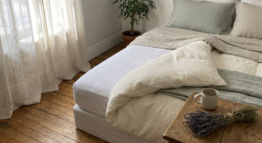 A beautifully made bed with a white mattress protector visible beneath a folded duvet, bathed in soft natural morning light in a warm, cozy bedroom.